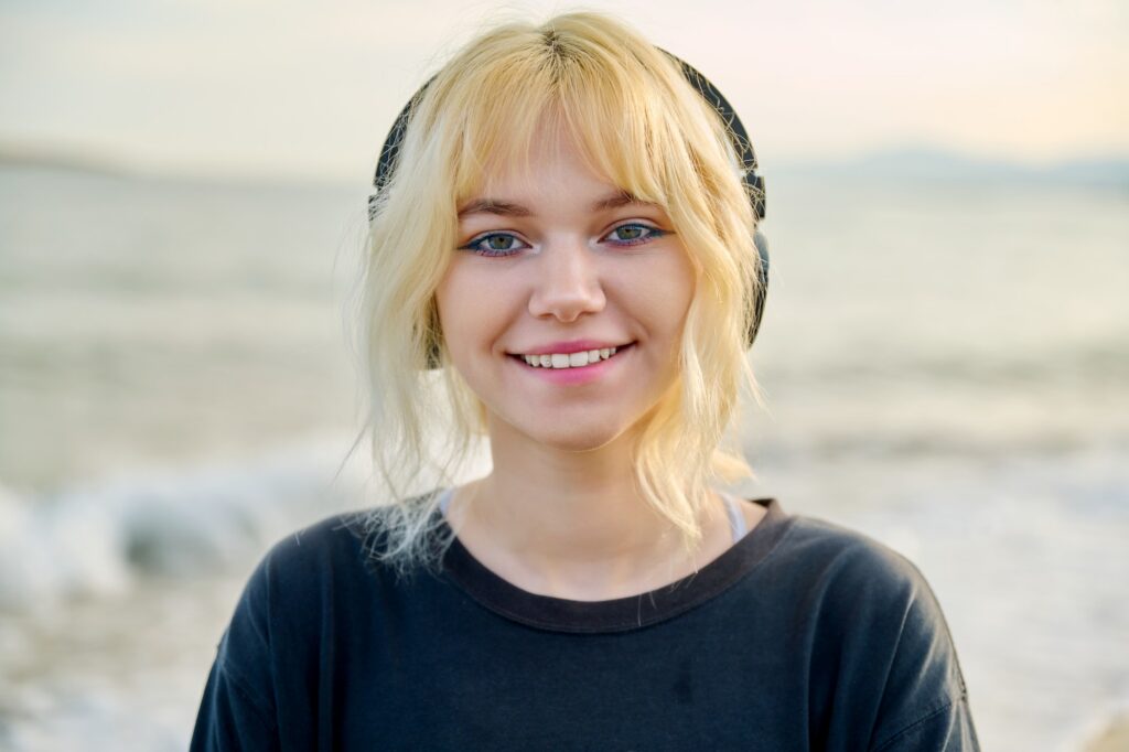 Portrait of smiling teenage female wearing headphones, on beach, close-up face, smile with teeth